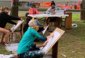 Child wearing mask sitting at easel and drawing outdoors. 