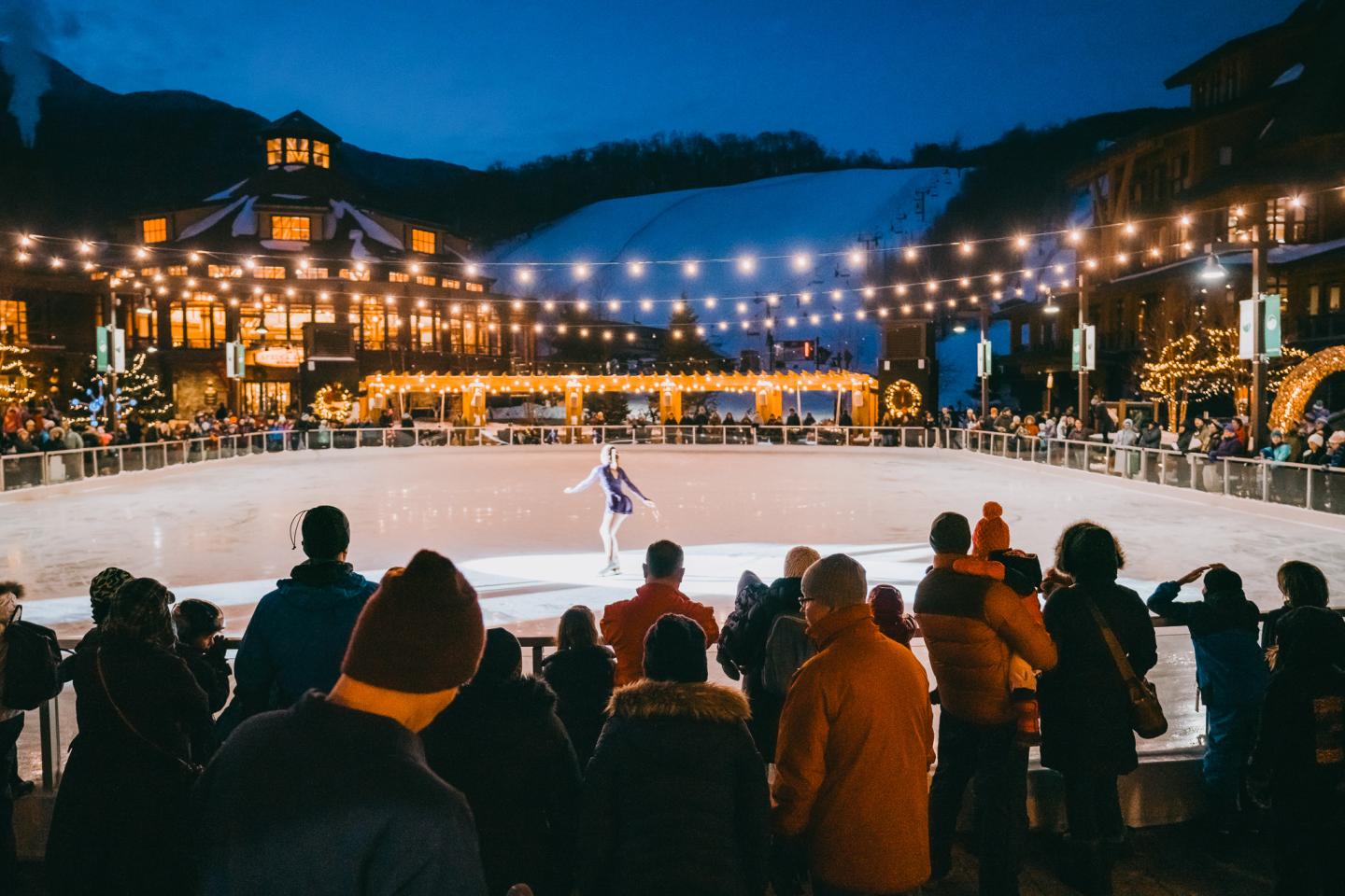 Crowd watching an ice skater perform on a lit outdoor rink at night.