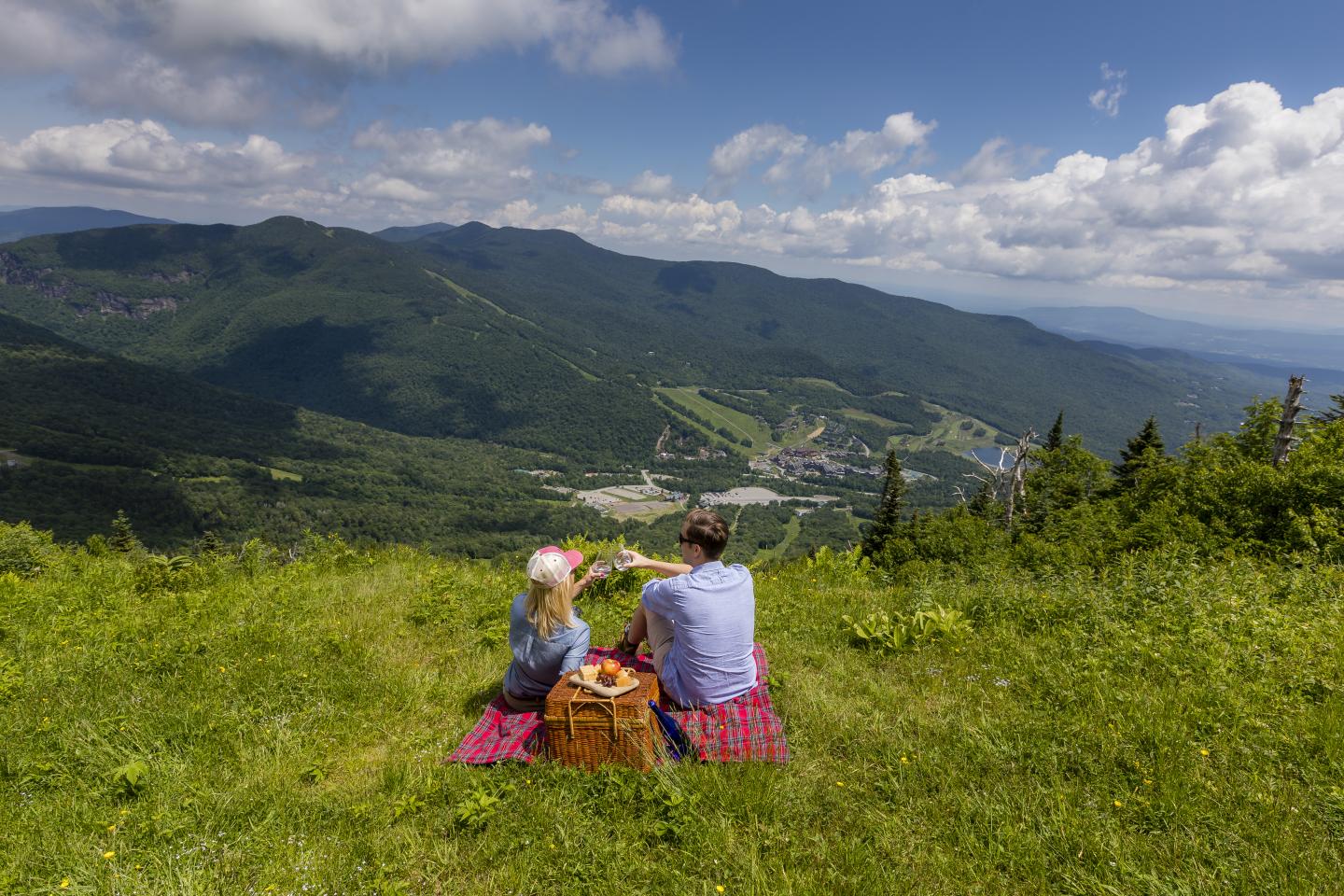 Two people having a picnic on a grassy hilltop overlooking mountains.