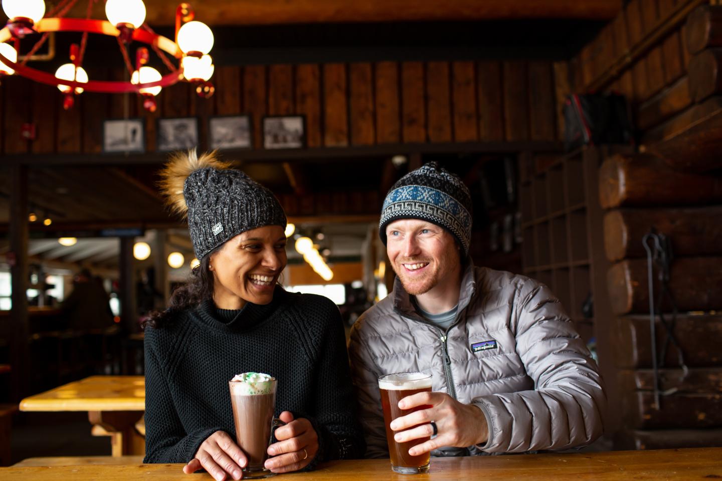 Cozy cafe setting with two people smiling, holding drinks, and wearing winter hats.