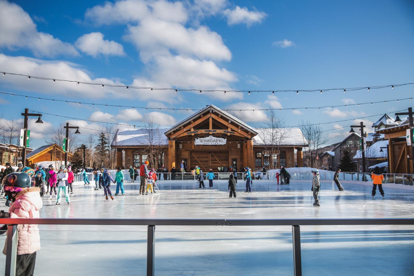 Skaters on an outdoor rink under a sunny, blue sky with clouds.