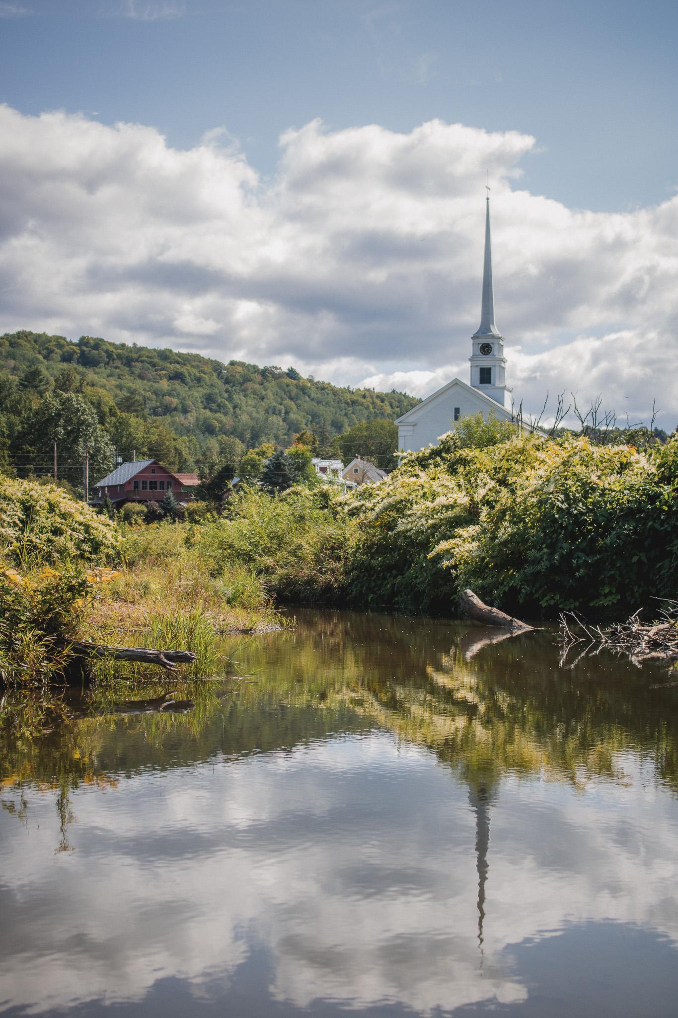 White church with tall steeple by a pond, surrounded by greenery under a cloudy sky.