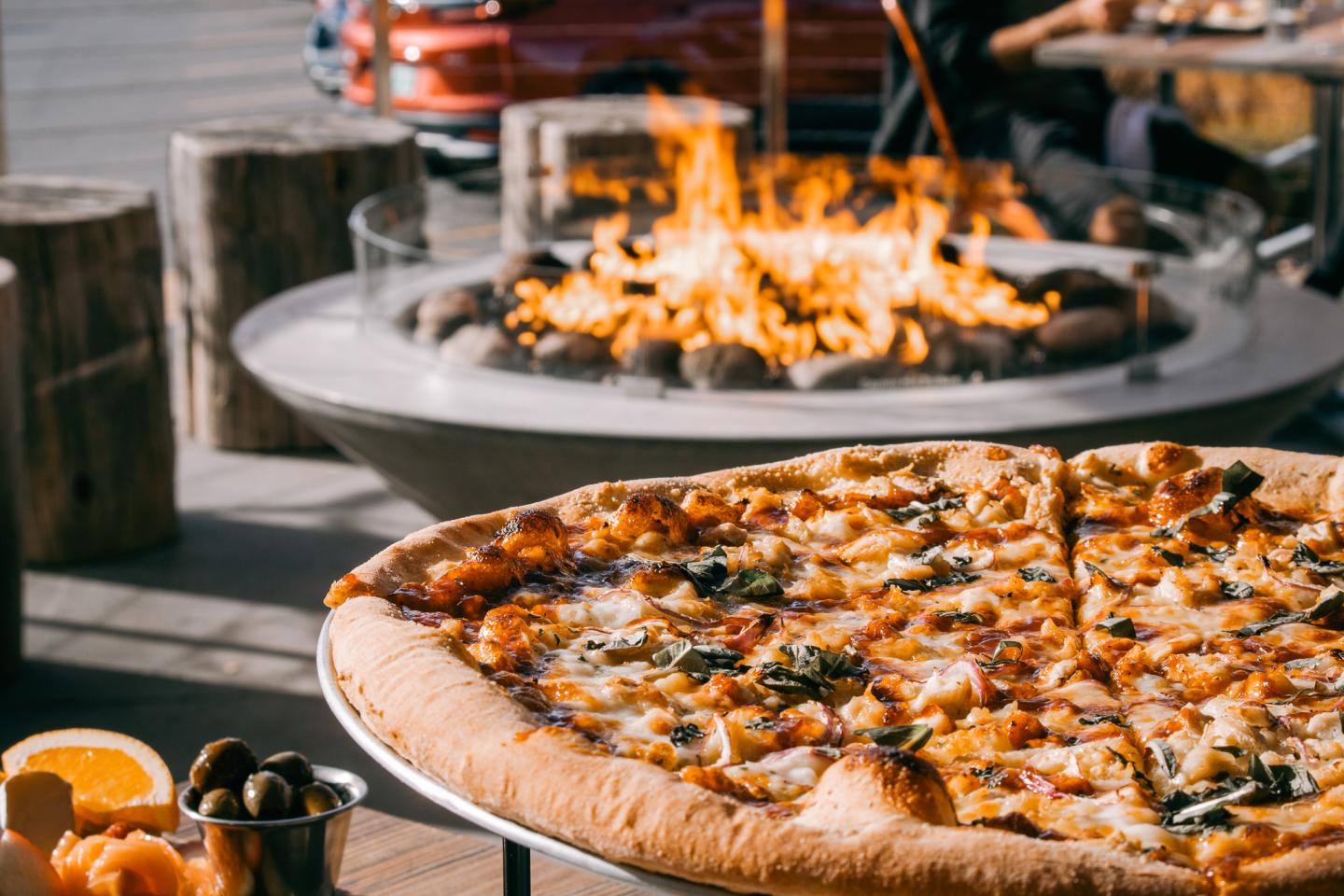 Pizza on table near outdoor fire pit with logs and a car in the background.
