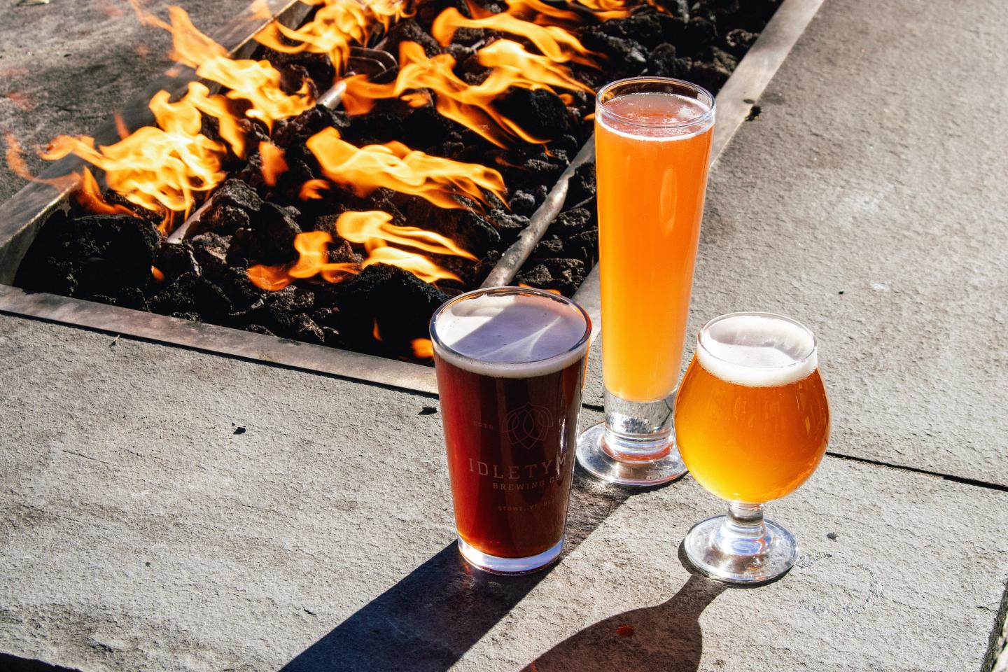 Three glasses of beer on a table near a flame-lit fire pit.