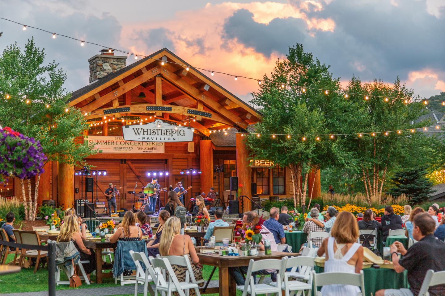 Outdoor concert with people seated at tables under string lights, sunset sky in background.