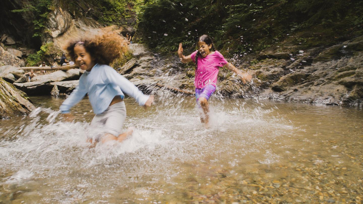 Children joyfully splashing in a shallow stream surrounded by trees.