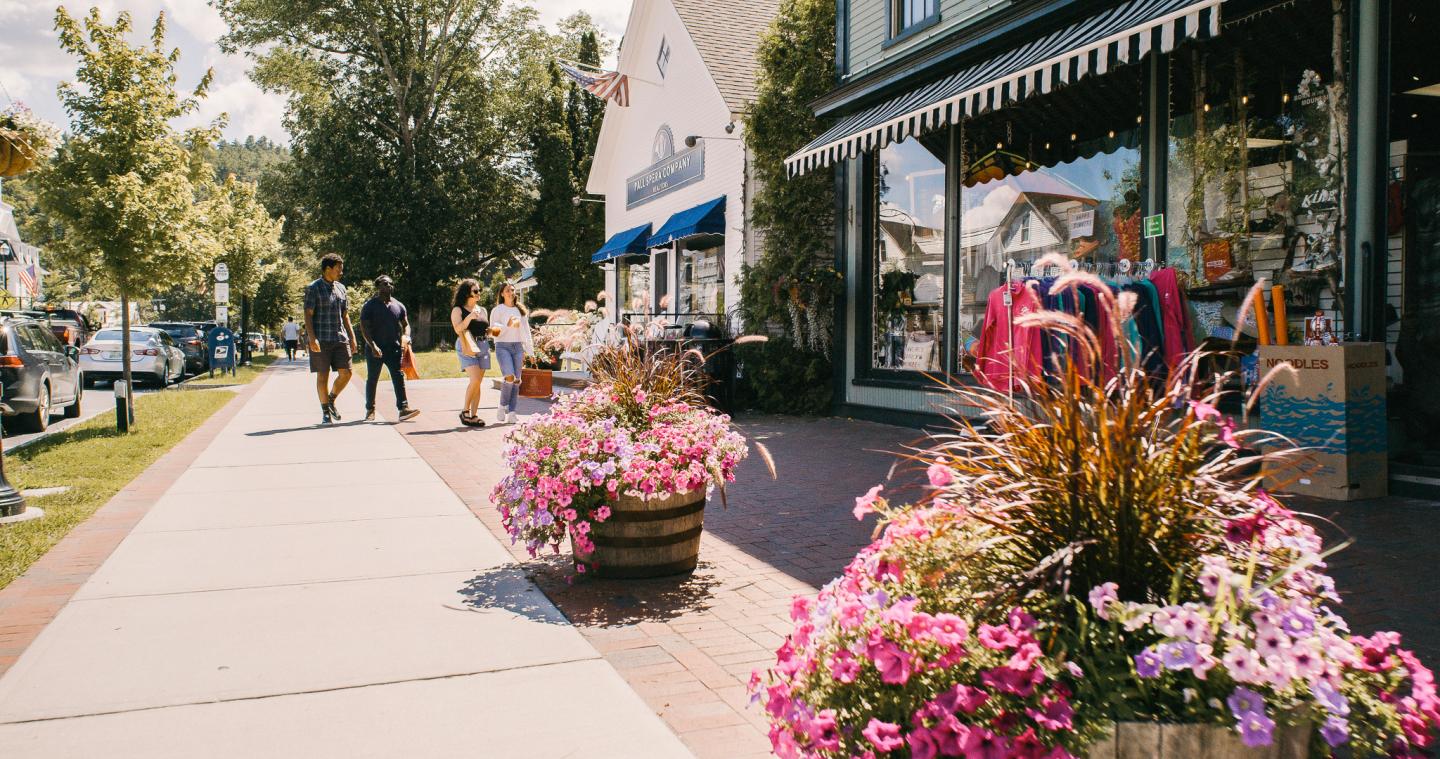 Sunny street with flower planters, storefronts, and people walking.