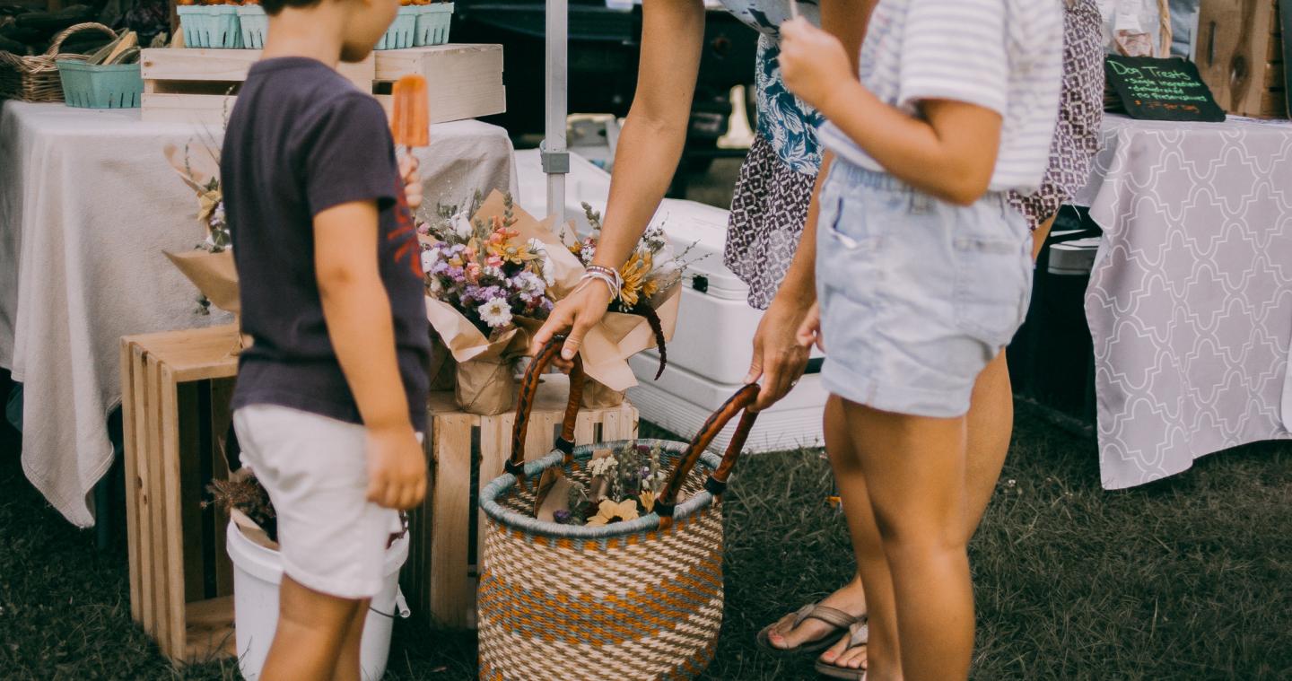 Market stall with baskets and flowers, two children and an adult browsing items.