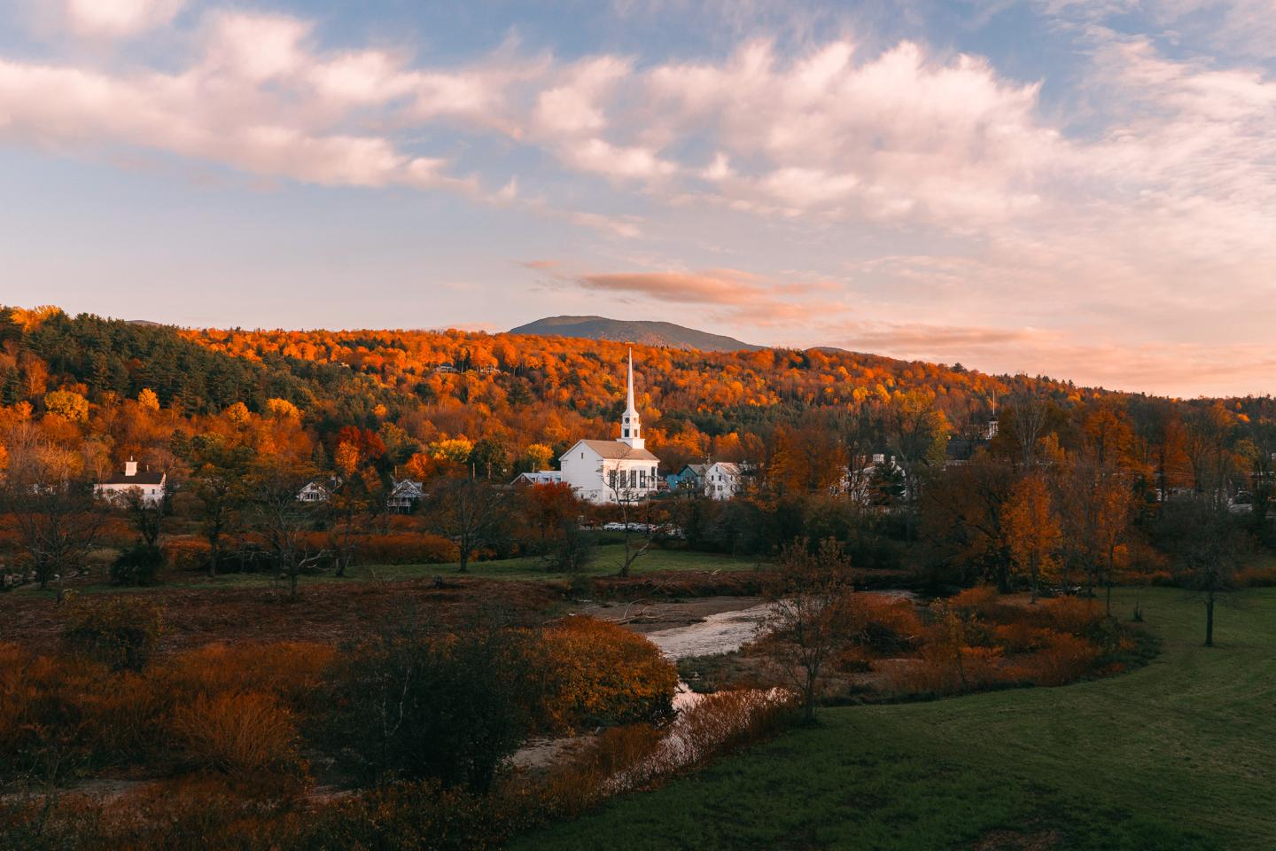 Autumn landscape with a white church, vibrant trees, and a mountain under a cloudy sky.