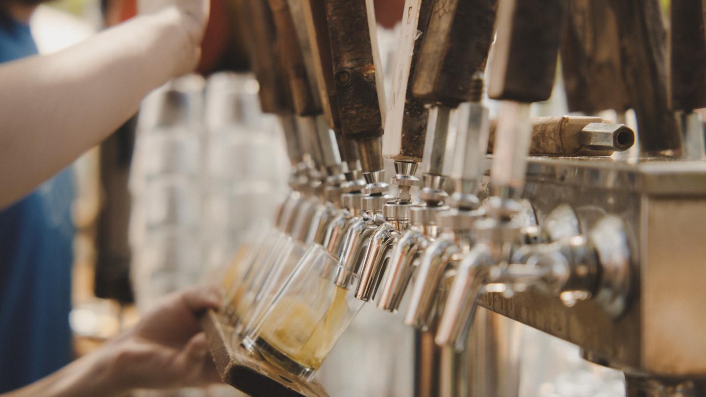 Hands pouring beer from multiple taps outdoors.