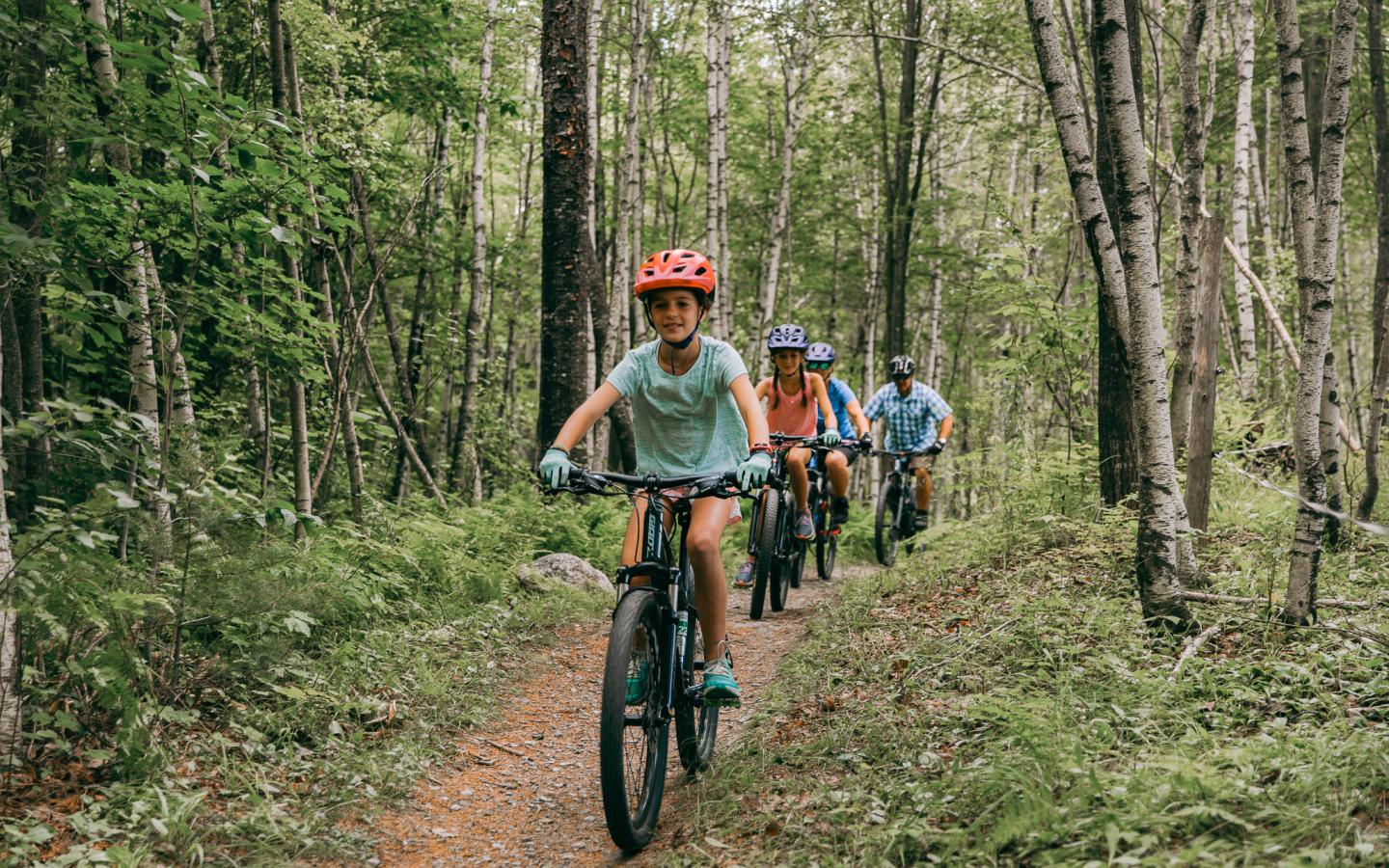 Cyclists riding on a forest trail, wearing helmets and casual clothing.