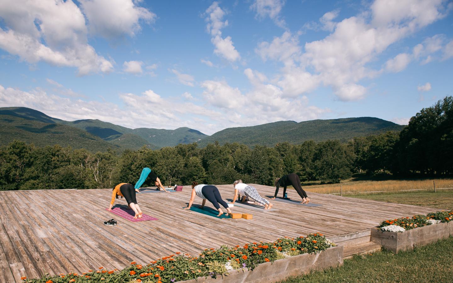 People doing yoga outdoors on a wooden platform with mountains in the background.