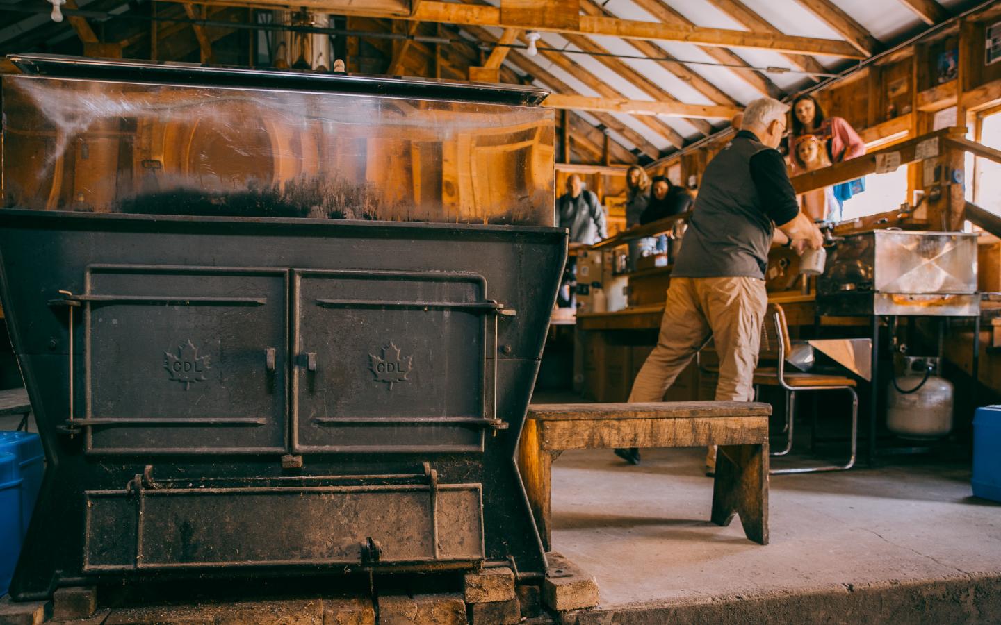 Wooden cabin interior with a large wood stove and a person tending to shelves.