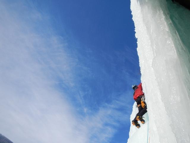 Climber ascending a steep ice wall under a clear blue sky.