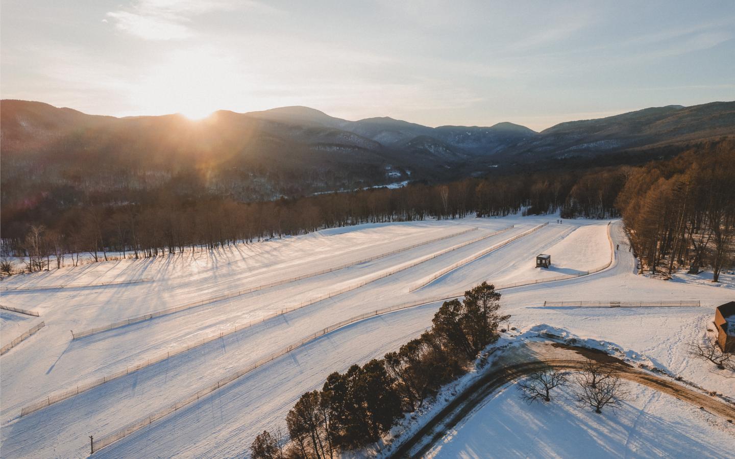 Snowy landscape at sunset, mountains and trees in the background.