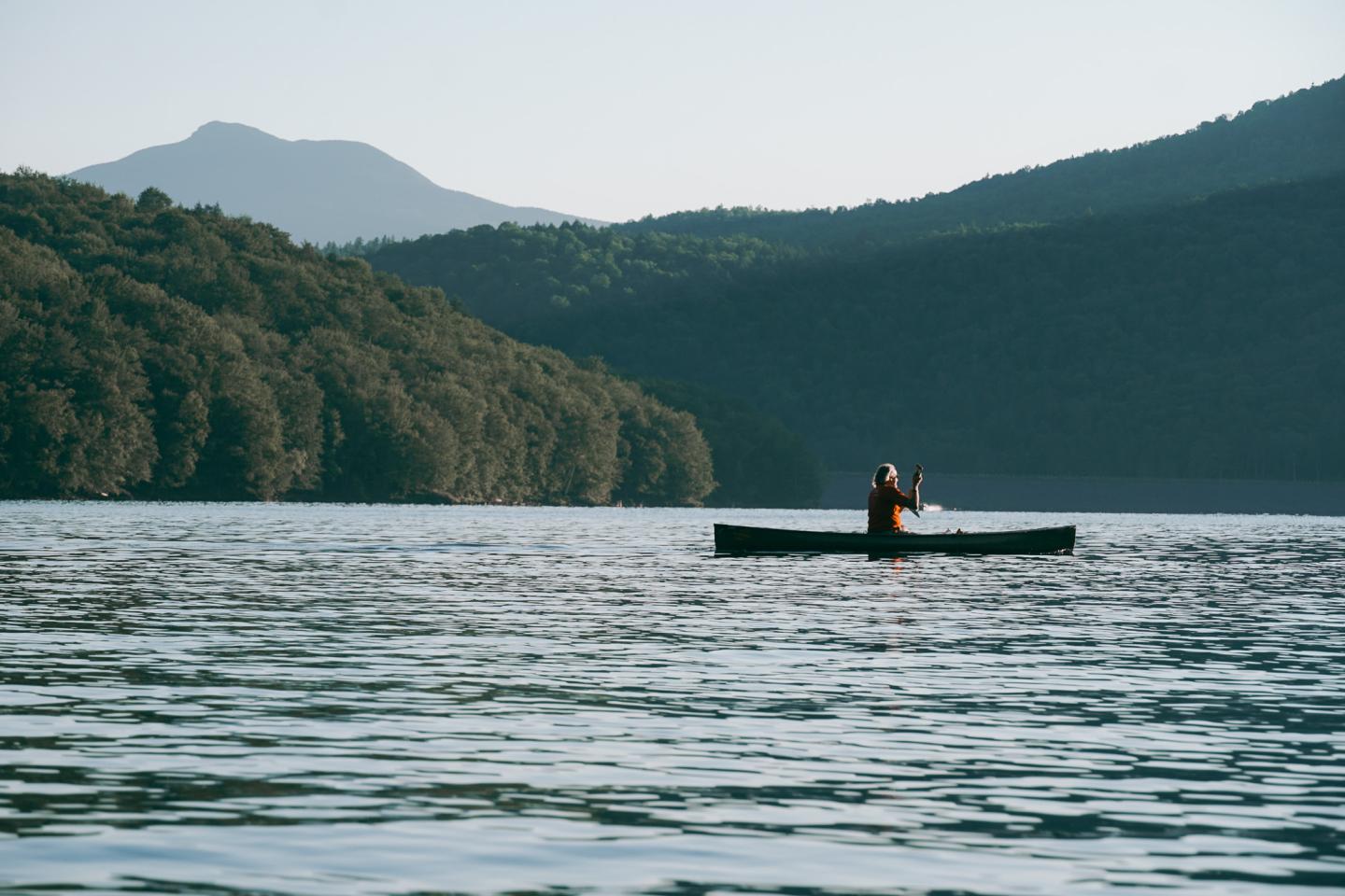 Kayaker on serene lake with forested hills and mountains in background.