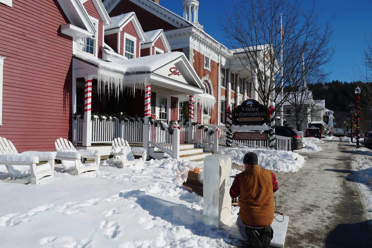 Man carving ice sculpture outside a red building on a snowy day.