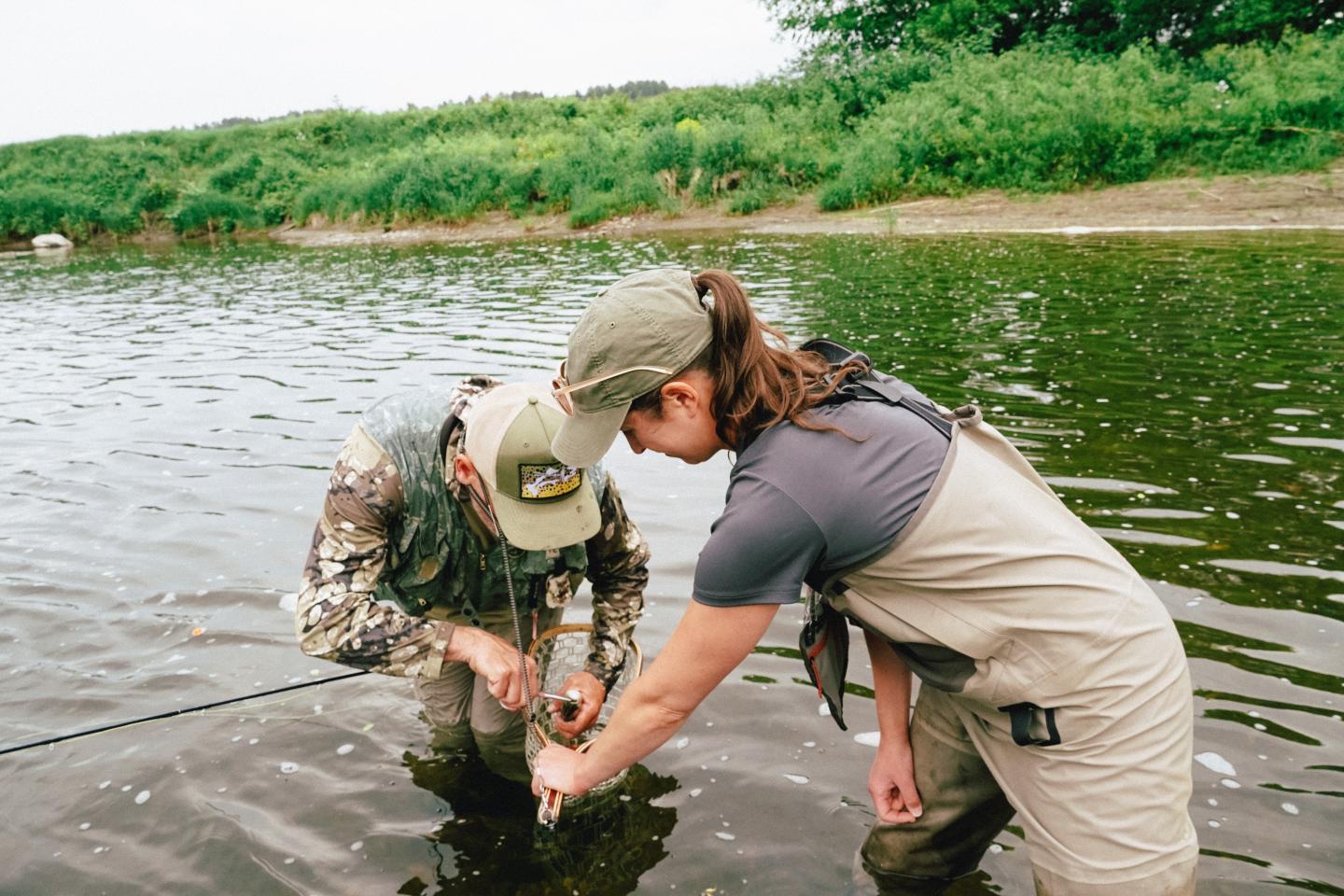 Two people fishing in a river, examining their catch.