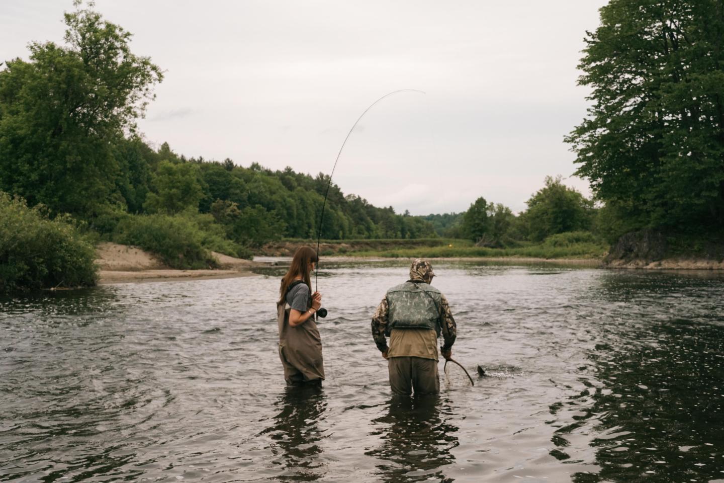 Two people fishing in a shallow river surrounded by trees.