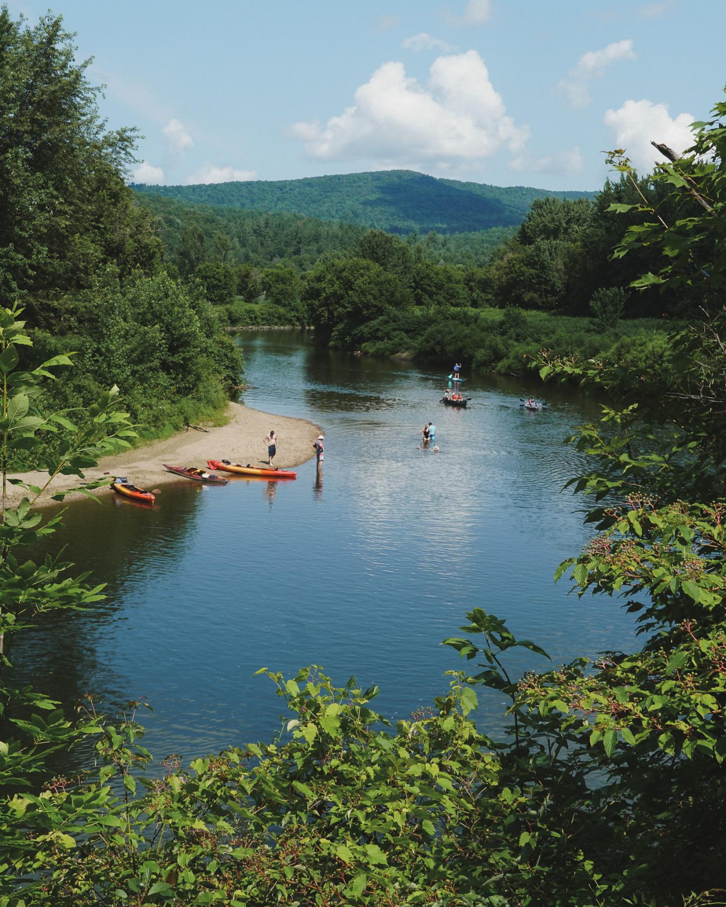 River with kayaks and swimmers, surrounded by lush greenery and distant hills.
