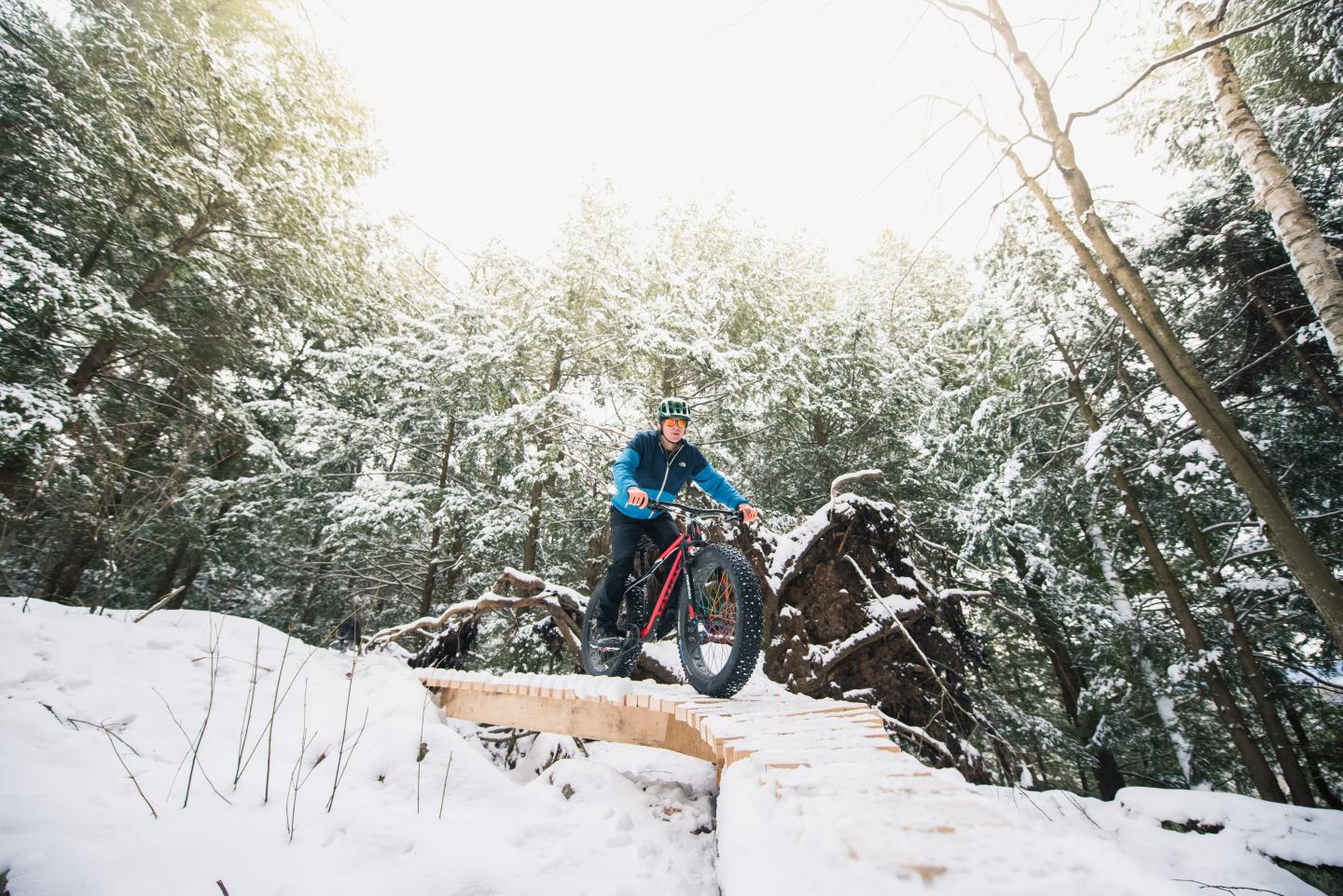 Cyclist on a snowy wooded trail, sunlight filtering through trees.