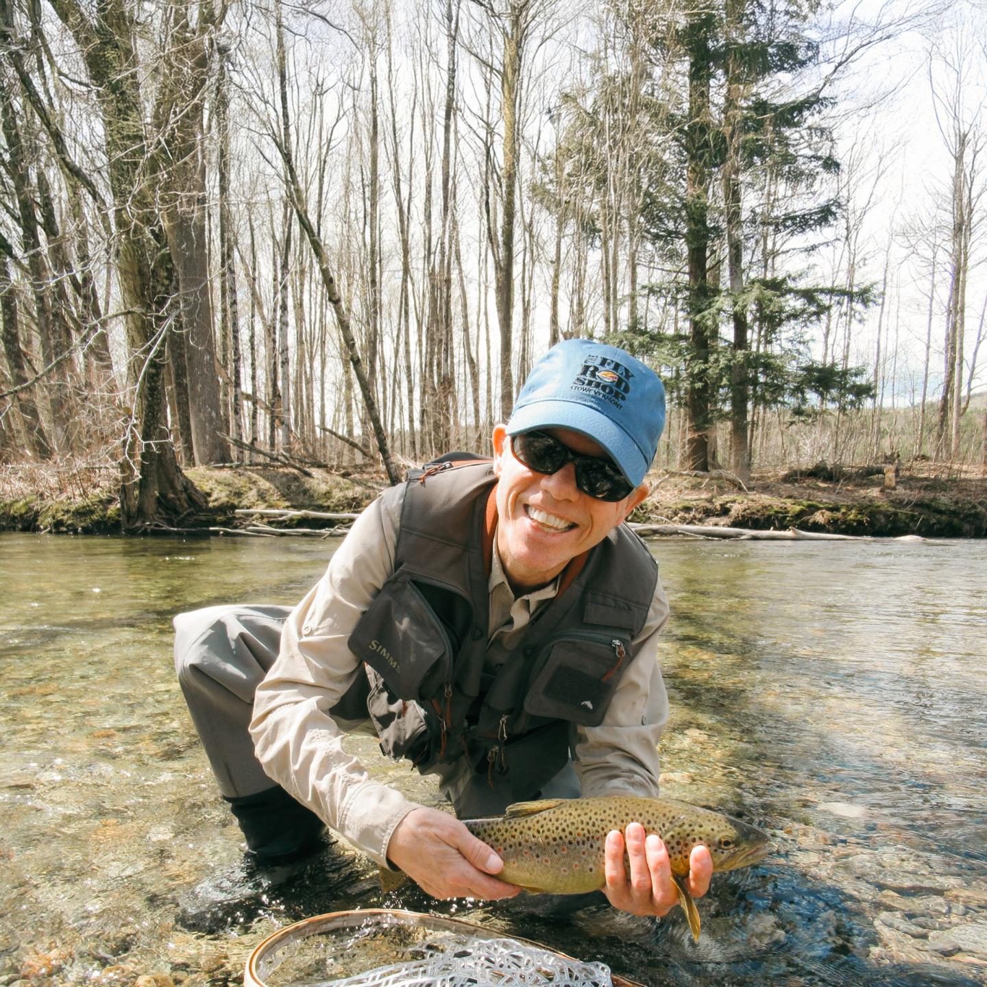 Man in a stream holding a fish, wearing sunglasses and a blue cap, surrounded by trees.