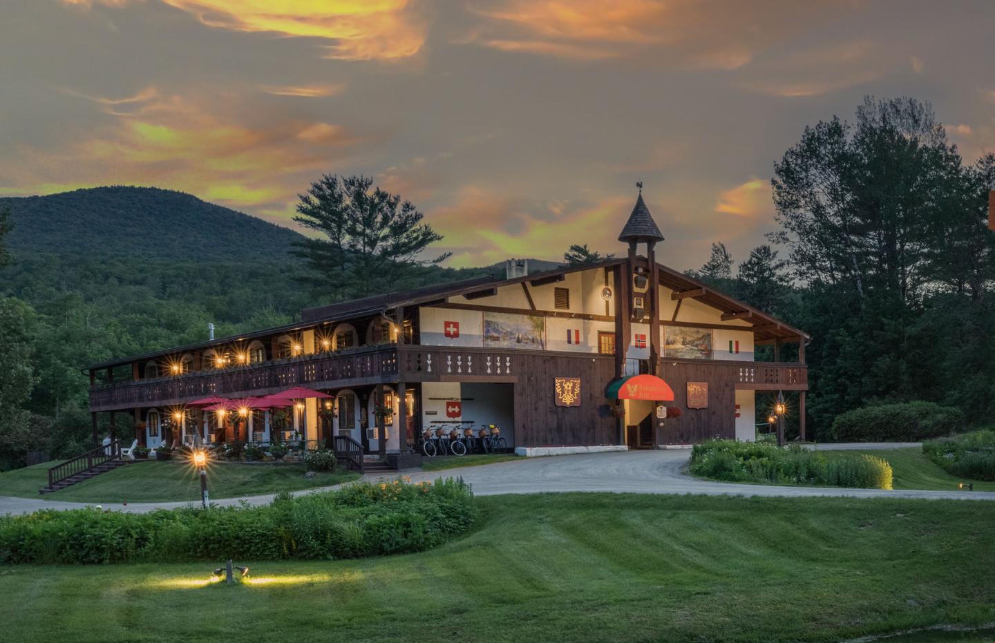 Wooden lodge with lights, surrounded by grass and trees, at sunset.