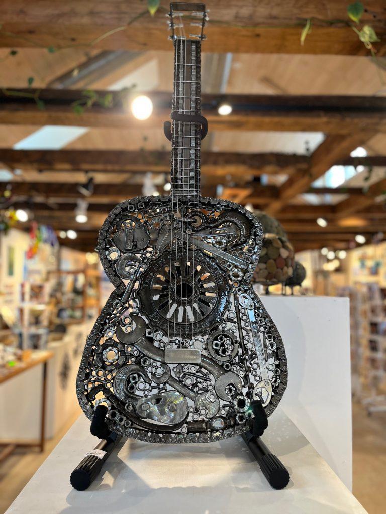 Steel sculpture of an ornate guitar on display in a wooden-ceiling room.