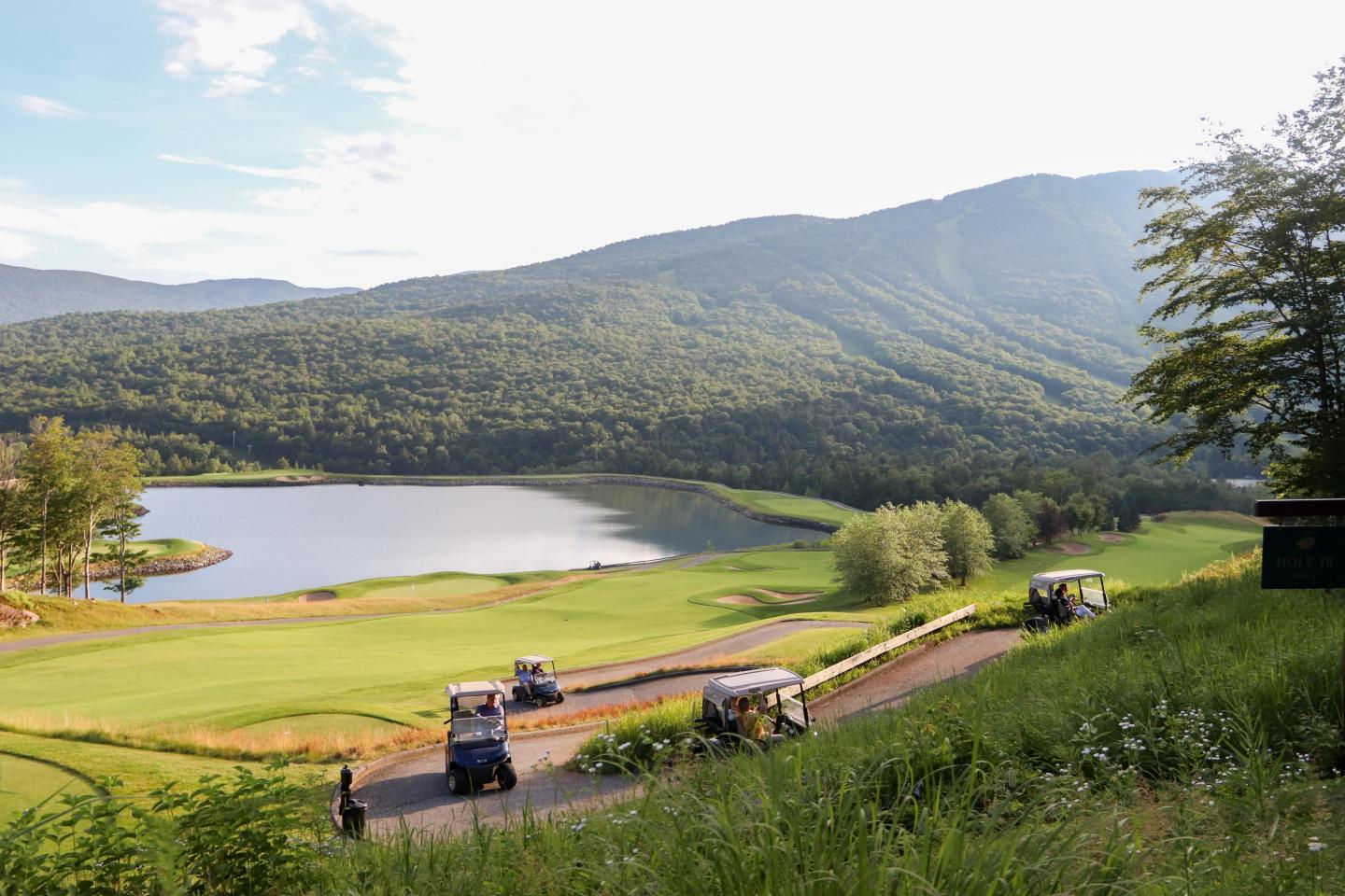 Golf course with carts, lake, and mountain backdrop.