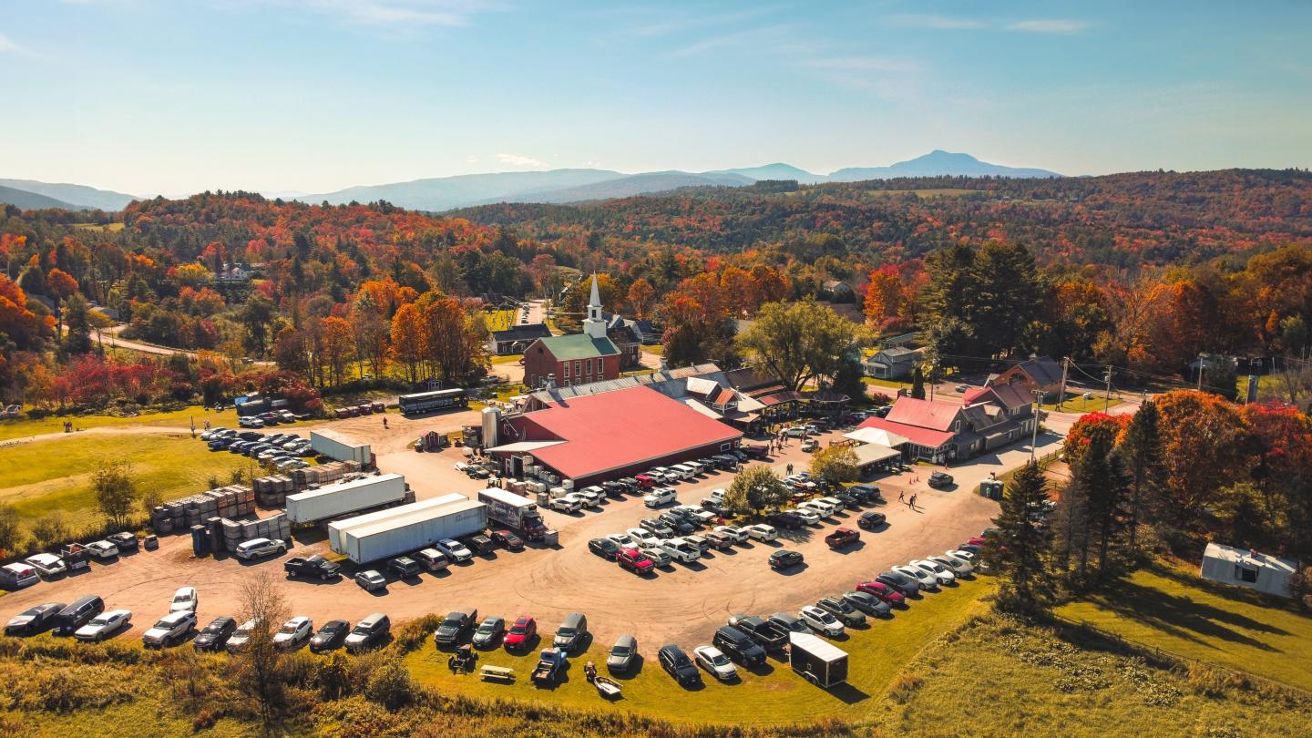Aerial view of a farm complex with red roofs, surrounded by autumn trees and parked cars.
