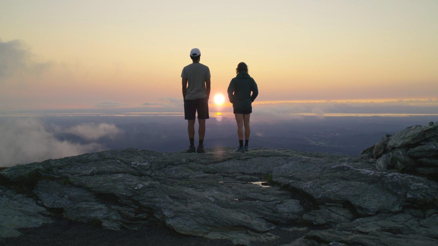 Two people standing on a rocky cliff, watching the sunset.