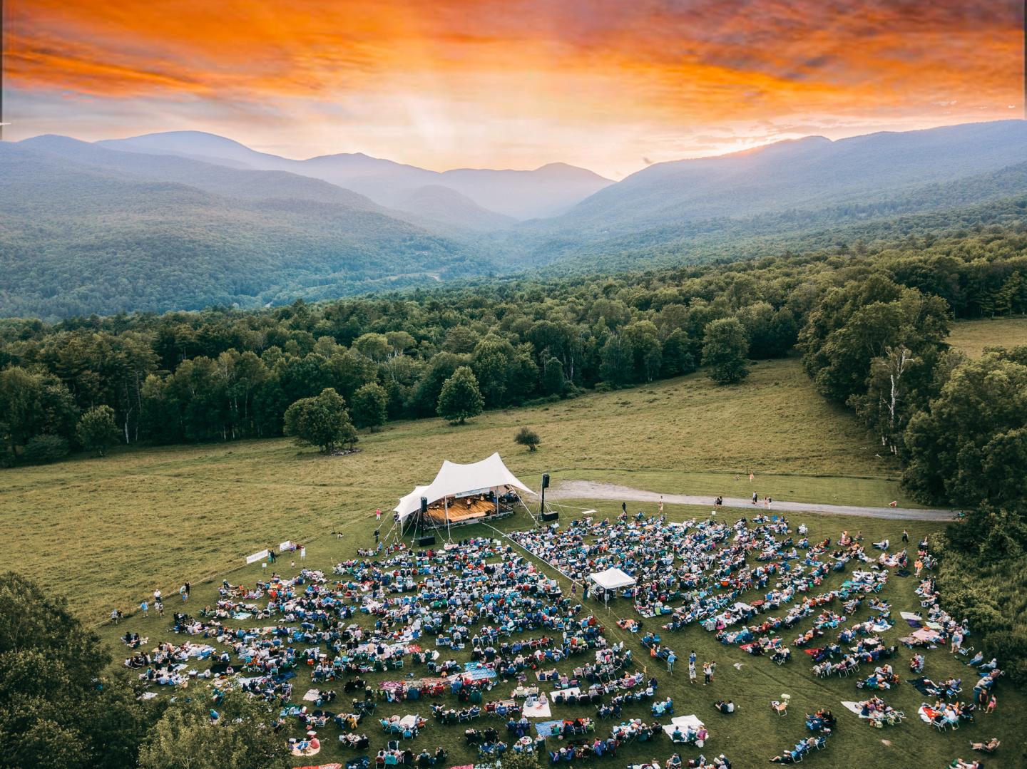 Outdoor concert with a large crowd, surrounded by green fields and mountains at sunset.