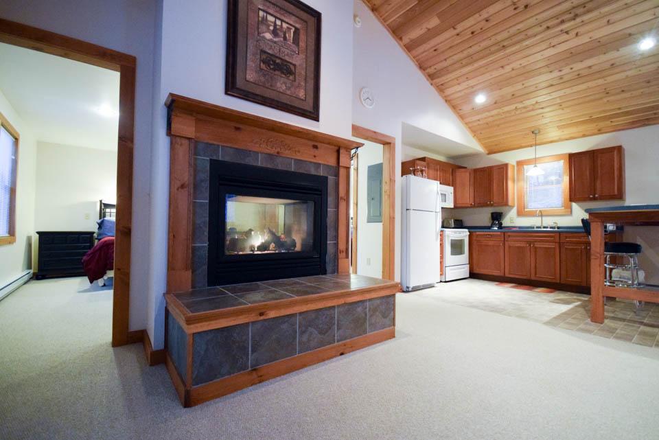 Cozy living room with fireplace, vaulted ceiling, and adjacent kitchen with wood cabinets.