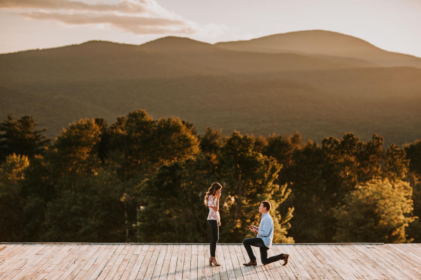 A man kneels in proposal at sunset on a wooden deck with mountains in the background.