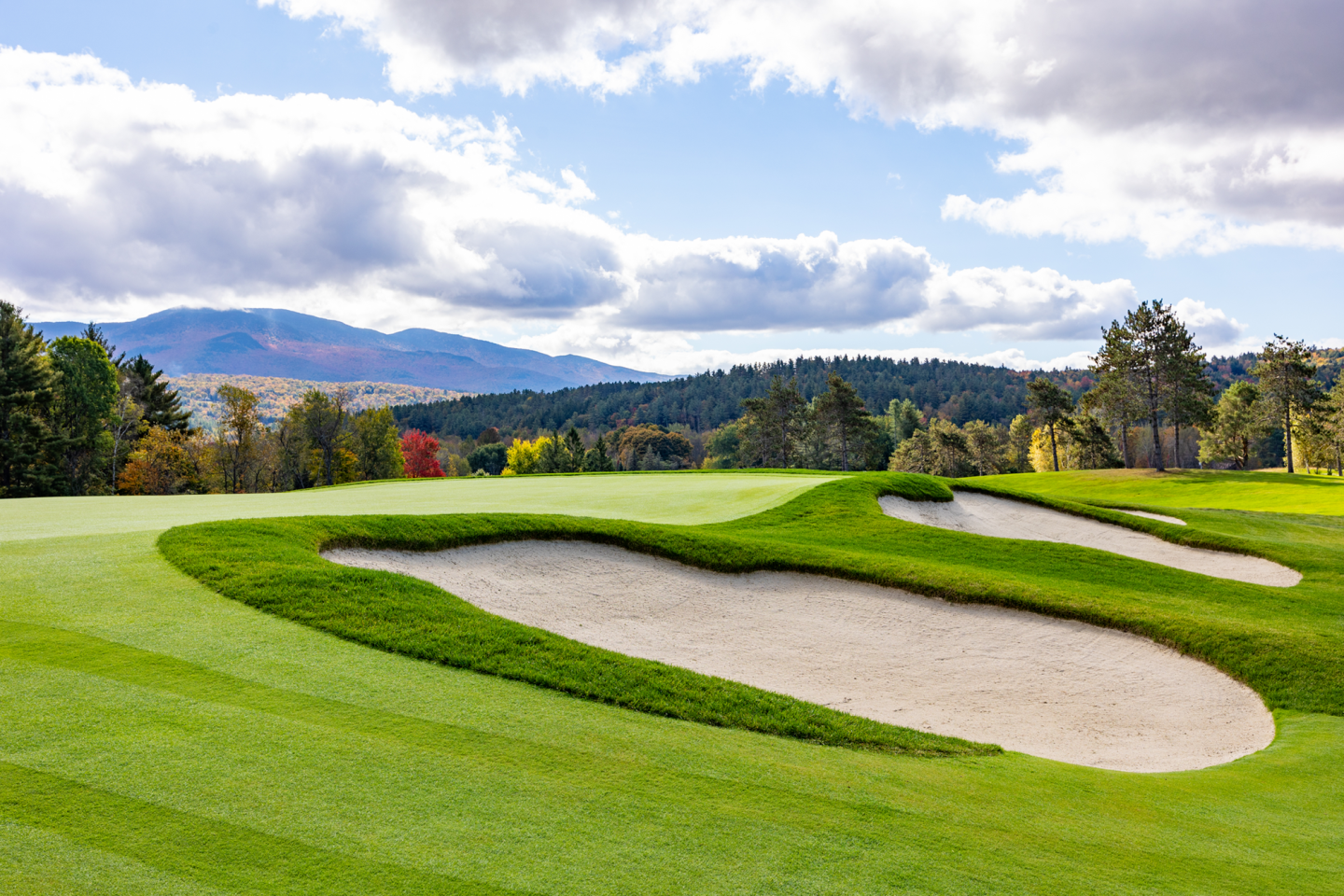Golf course with sand bunkers, green grass, mountains, and cloudy sky.