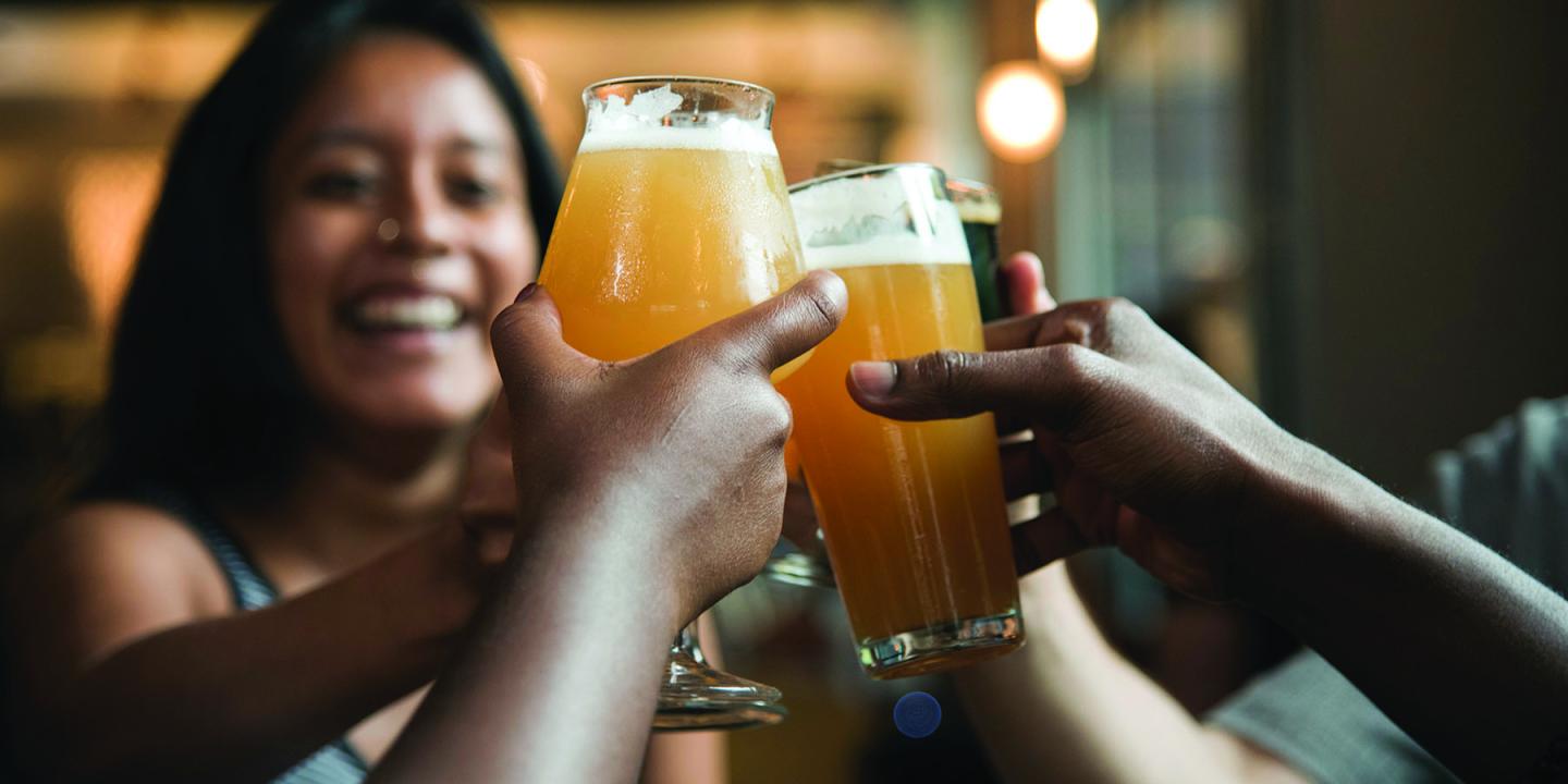 Cheers with glasses of beer, smiling woman in the background.