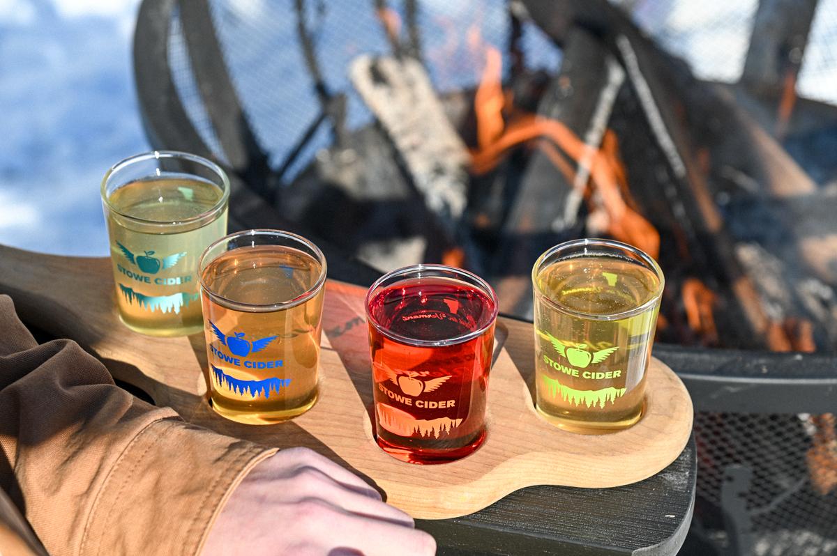 Four colorful drinks on a wooden board near an outdoor fire pit.