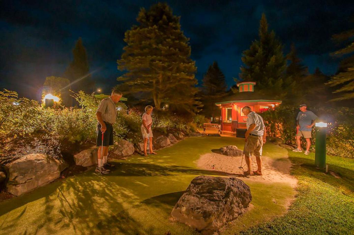 Night mini-golf game with four people, surrounded by lit trees and rocks.
