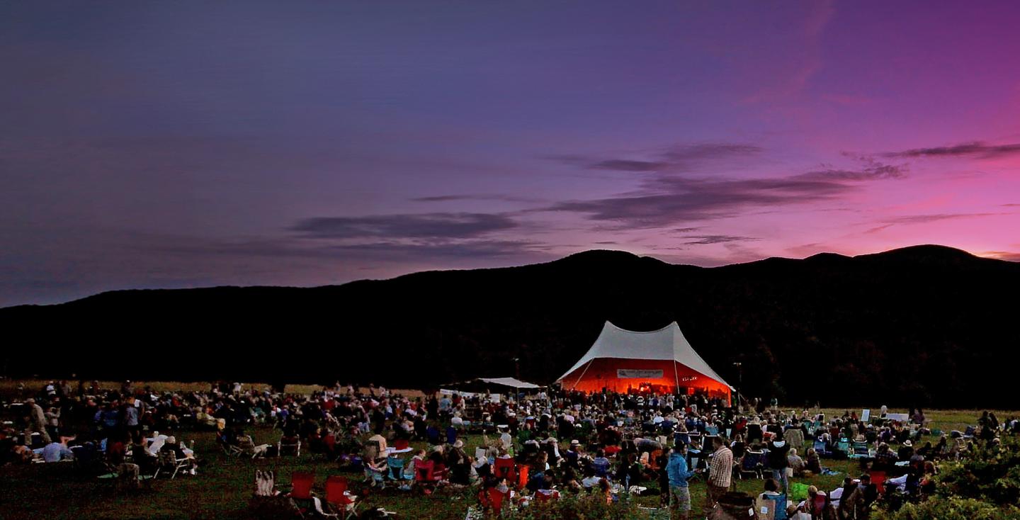 Outdoor concert at sunset with a large crowd and mountains in the background.