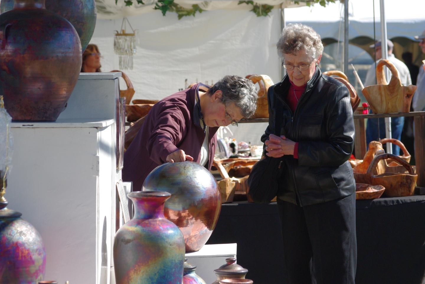 Two people browse pottery at an outdoor market stall.