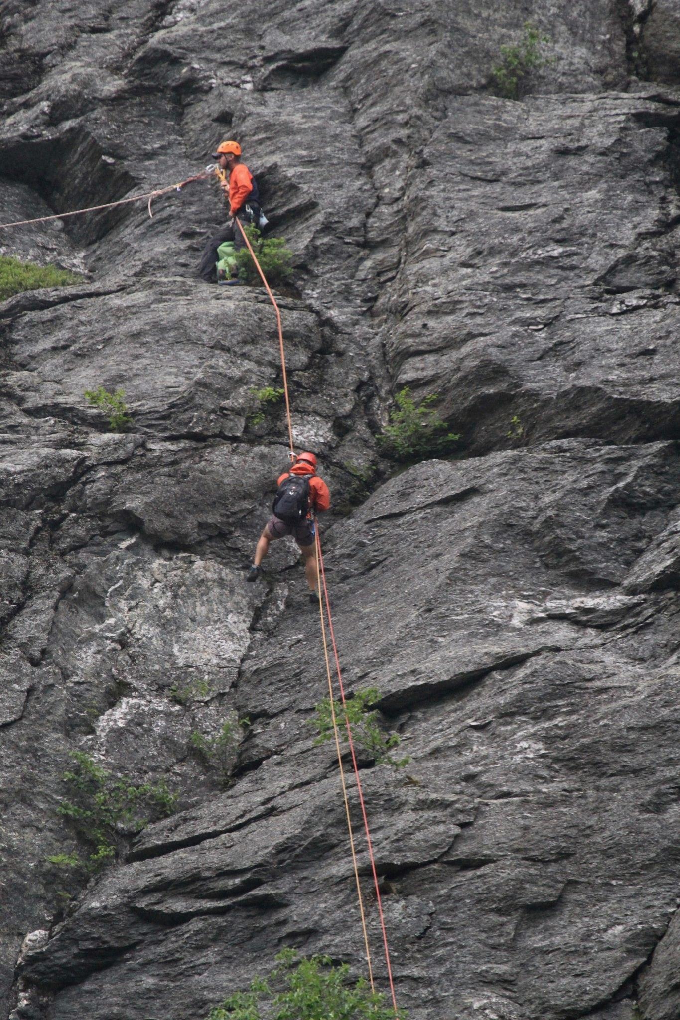 Rock climbers in orange helmets scaling a steep gray cliff.