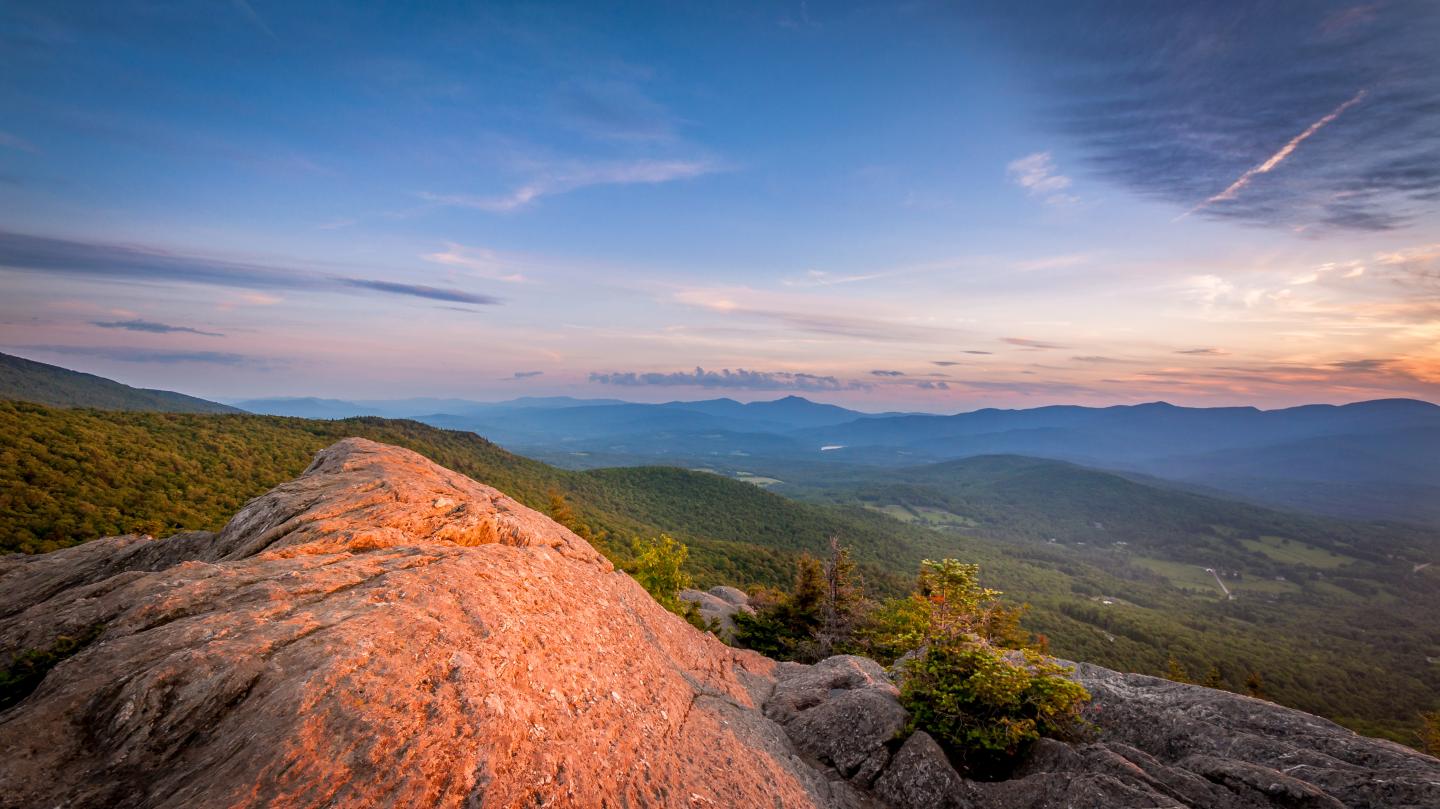 Sunset over a rocky ledge with a forested valley below.