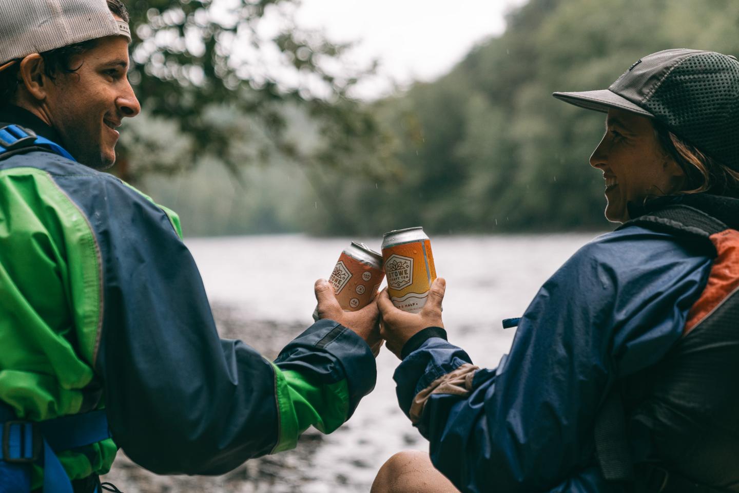 Two people in rain jackets cheer with cans by a river.