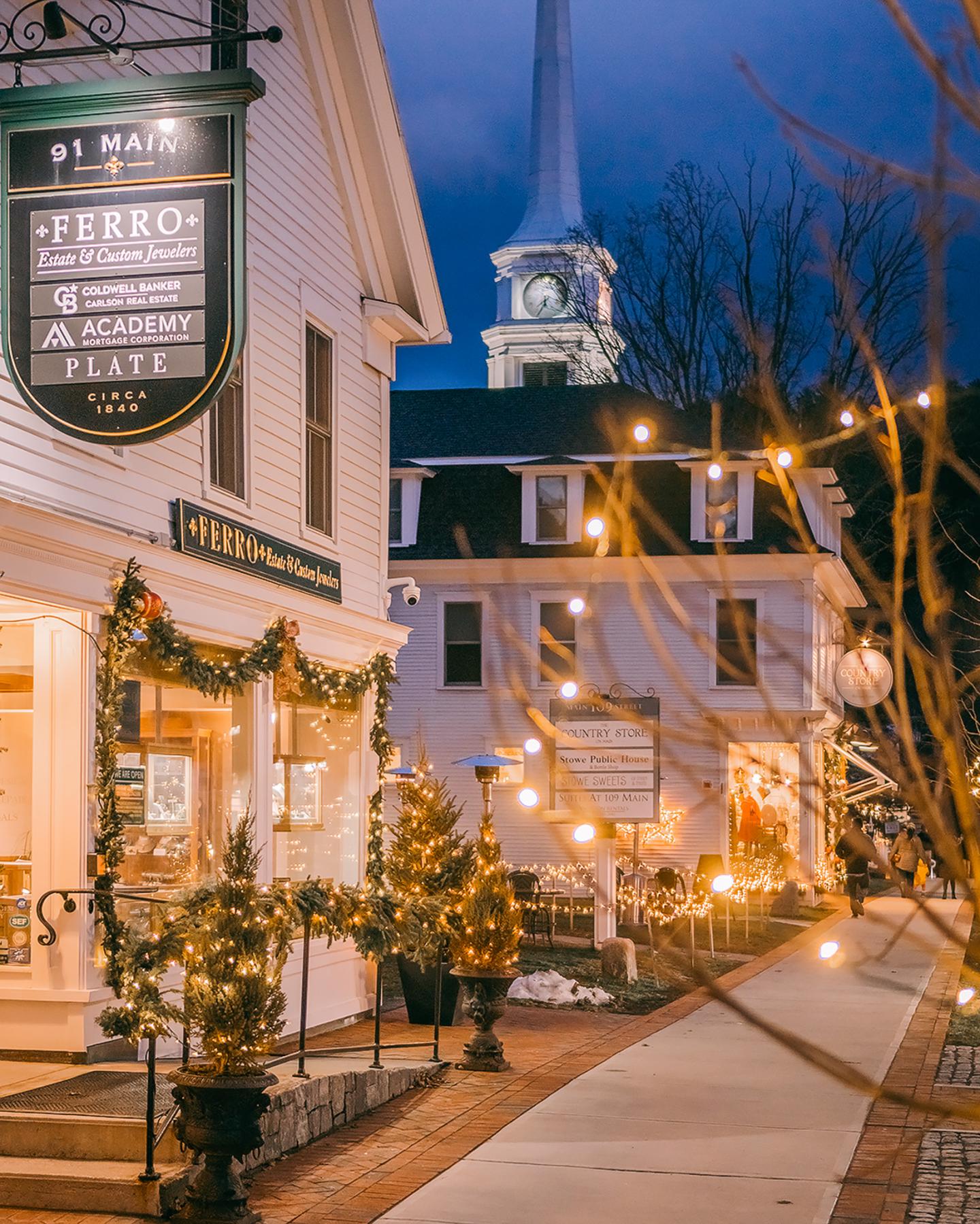 Festive street with lights, decorated shops, and a church steeple at dusk.