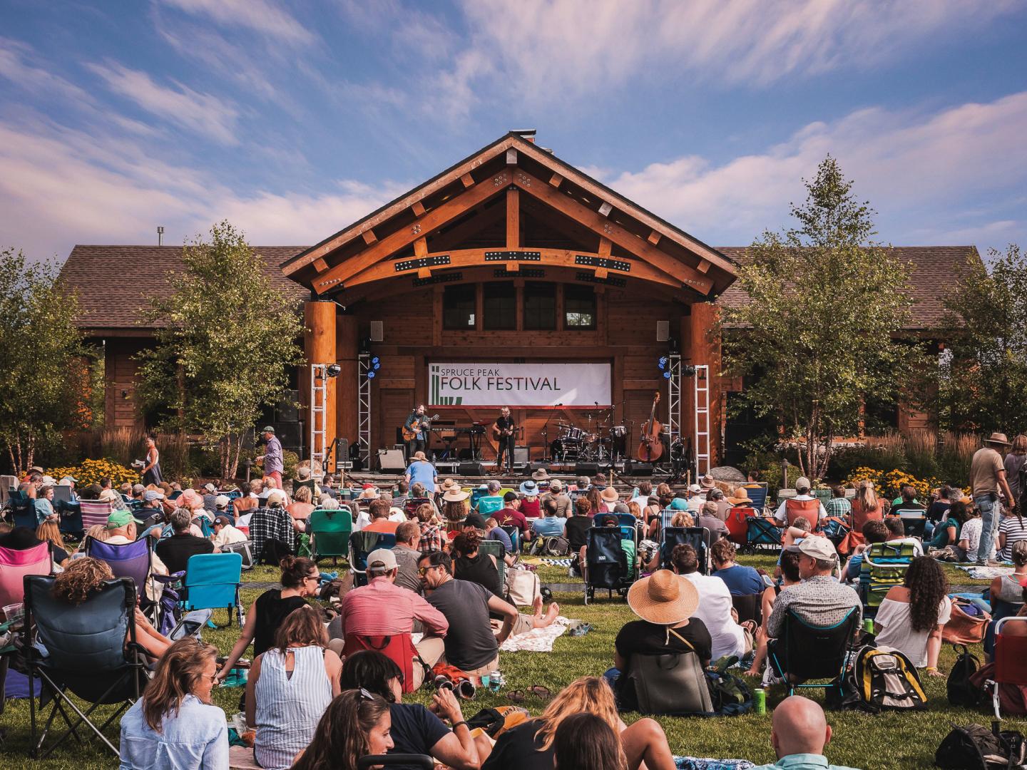 Outdoor concert with a large crowd sitting on grass in front of a wooden stage.
