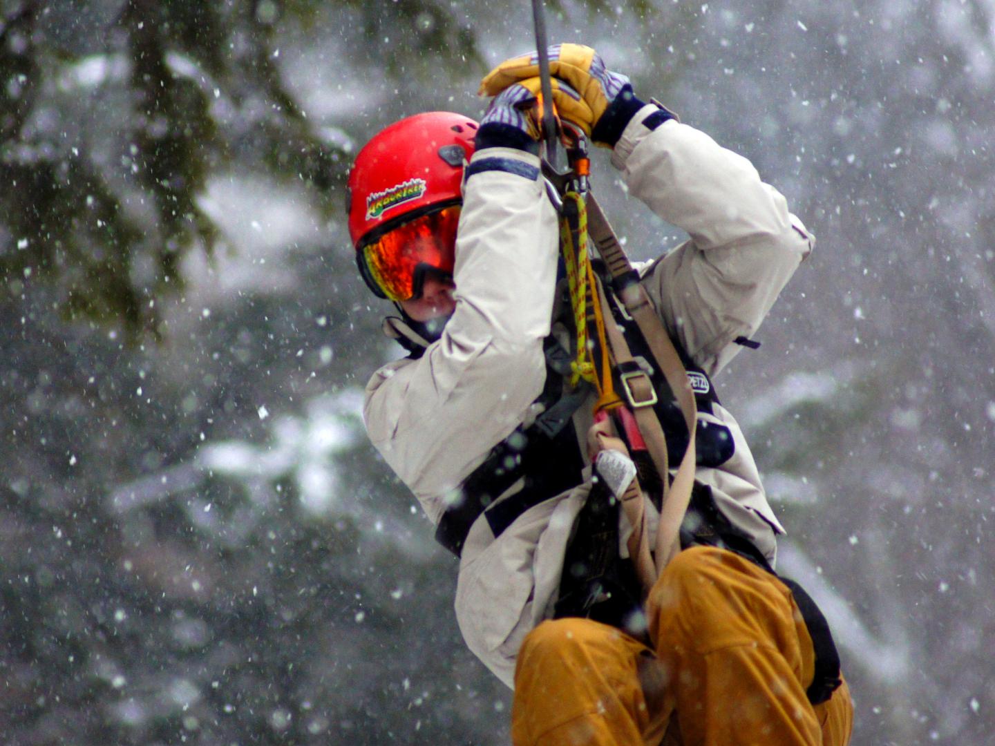 Person zip-lining in snow, wearing a red helmet and beige coat.