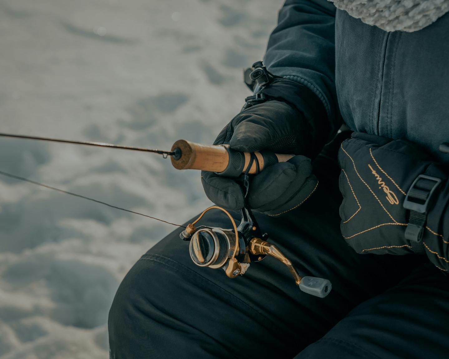Person ice fishing with a rod and reel, wearing gloves and winter clothing.