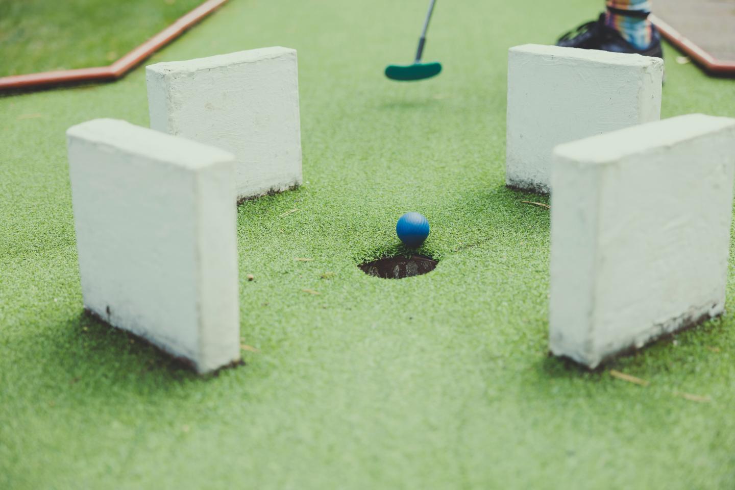 Mini-golf green with blue ball near a hole, surrounded by white barriers.