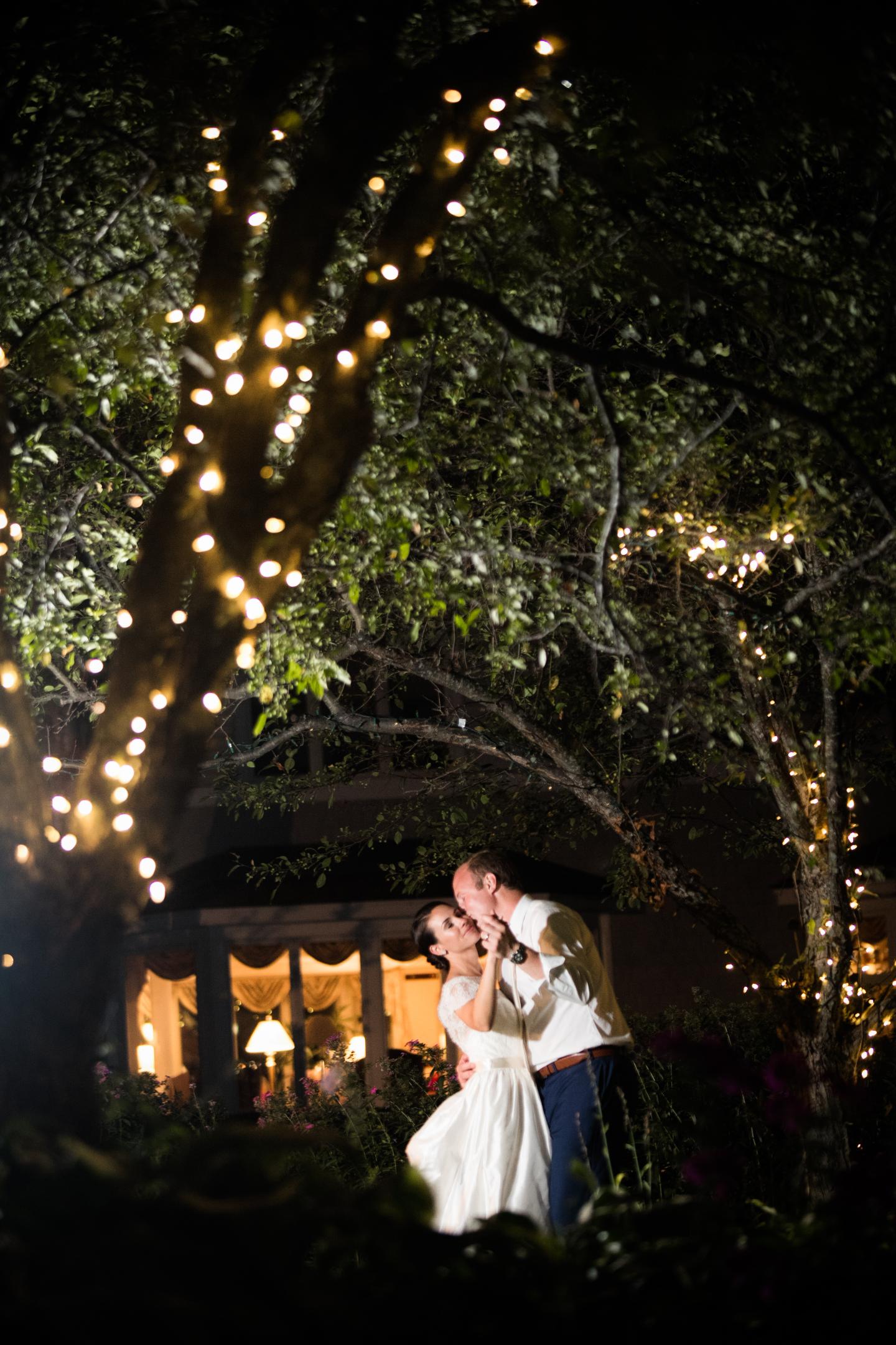 Bride and groom dancing under tree with string lights at night.