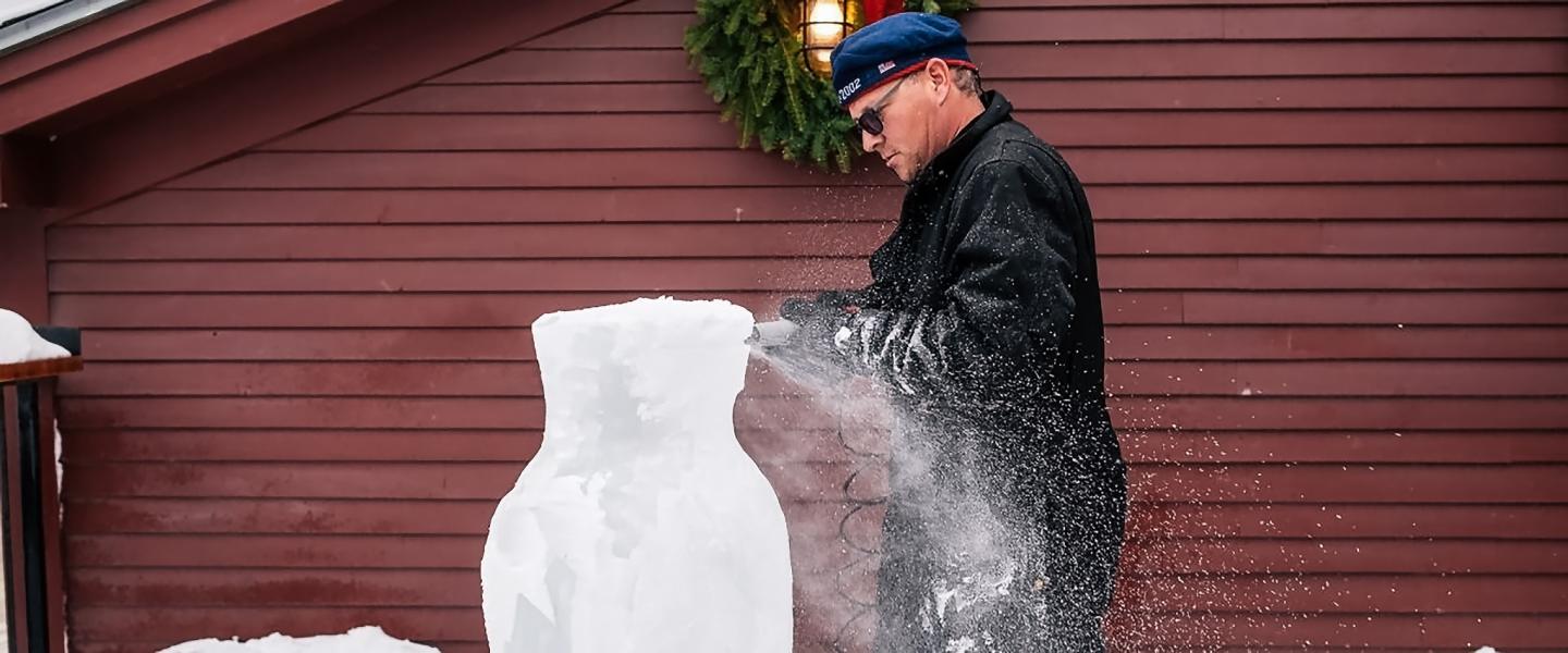 Man carving an ice sculpture in front of a red wooden building.
